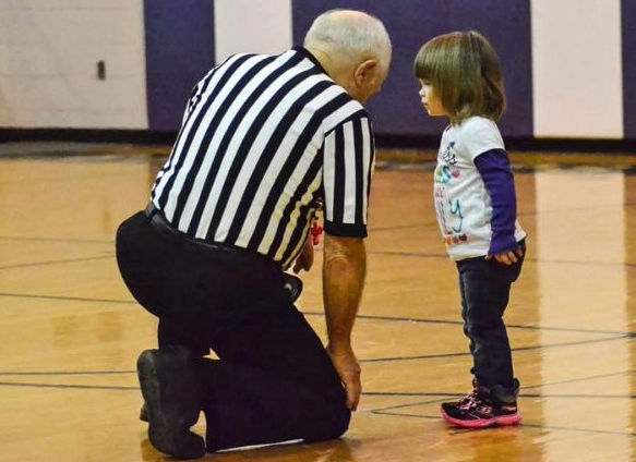 Basketball referree Paul Cooley shown down on a knee listening to small young girl on the court.